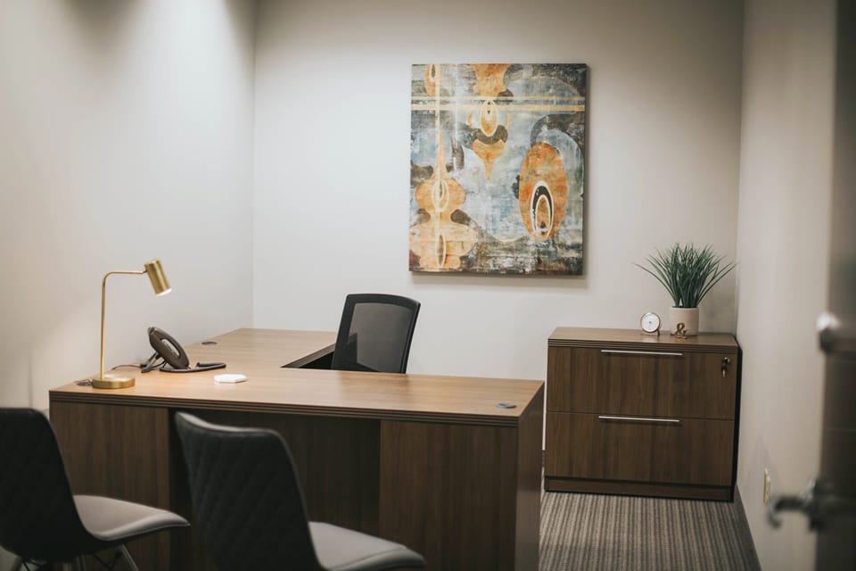 Private office with L-shaped desk, rolling desk chair, guest seating, lighting and artworks, phone access, and a filing cabinet